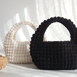 K&ouml;nnte beinhalten: Zwei geh&auml;kelte Handtaschen, eine wei&szlig;e und eine schwarze, mit abgerundeter Form und einem einzigen Henkel. Die Taschen sind mit einem strukturierten, bauschigen Stich gearbeitet.