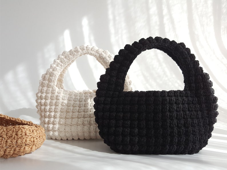 K&ouml;nnte beinhalten: Zwei geh&auml;kelte Handtaschen, eine wei&szlig;e und eine schwarze, mit abgerundeter Form und einem einzigen Henkel. Die Taschen sind mit einem strukturierten, bauschigen Stich gearbeitet.