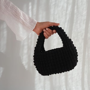 K&ouml;nnte beinhalten: Eine schwarze, geh&auml;kelte Handtasche mit abgerundeter Form und einem einzigen Henkel.