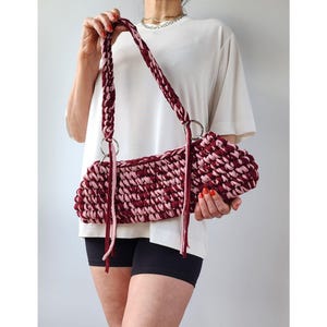 May include: A burgundy and pink crocheted handbag with silver metal rings and a braided strap. The bag has a rectangular shape and is held by a person. The bag's color scheme is a mix of burgundy and pink, creating a textured appearance.