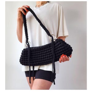 May include: A black, crocheted shoulder bag with a rectangular shape. The bag features a thick, braided strap and is attached to silver-colored rings. Long, thin tassels hang from the bag's sides. The bag is held by a person wearing a white shirt and black shorts.