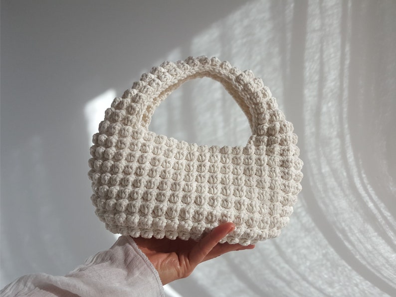 K&ouml;nnte beinhalten: Eine wei&szlig;e geh&auml;kelte Handtasche mit abgerundeter Form und einem einzigen Henkel. Die Tasche ist mit einem strukturierten Stich gearbeitet, der eine unebene Oberfl&auml;che erzeugt.