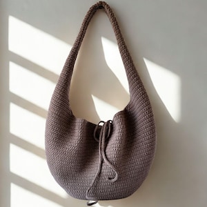 Puede incluir: Un bolso de hombro de ganchillo en color malva apagado, colgado contra una pared neutra. El bolso presenta una forma redondeada de media luna con una correa larga e integrada. Un cierre de cordón es visible en la parte superior. La textura del bolso está definida por las puntadas de ganchillo visibles.