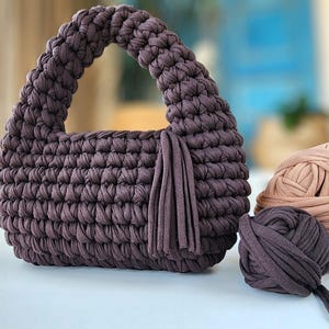 以下が含まれることがあります： 編み込みのハンドルとタッセルが付いた、手作りの濃い紫色のかぎ針編みハンドバッグ。バッグの横には、明るい茶色と濃い紫色の2つの毛糸玉があり、手芸プロジェクトを示唆しています。