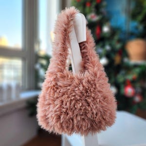 Puede incluir: Un bolso de mano de crochet rosa y esponjoso con una correa larga.