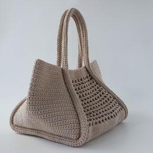 Op de afbeelding: Een beige gehaakte tote bag met een geweven patroon aan de ene kant. De tas heeft twee dikke gehaakte handvatten.