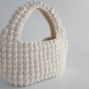 K&ouml;nnte beinhalten: Eine wei&szlig;e geh&auml;kelte Handtasche mit abgerundeter Form und einem einzigen Henkel. Die Tasche ist mit einem strukturierten Stich gearbeitet, der eine unebene Oberfl&auml;che erzeugt.