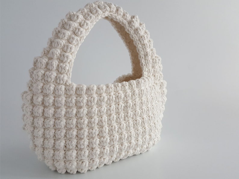 K&ouml;nnte beinhalten: Eine wei&szlig;e geh&auml;kelte Handtasche mit abgerundeter Form und einem einzigen Henkel. Die Tasche ist mit einem strukturierten Stich gearbeitet, der eine unebene Oberfl&auml;che erzeugt.