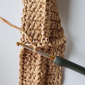 May include: Close-up of a crochet project in progress. A beige woven material is being worked with a gold-colored crochet hook with a dark green handle. The hook is inserted into a loop of the woven material.