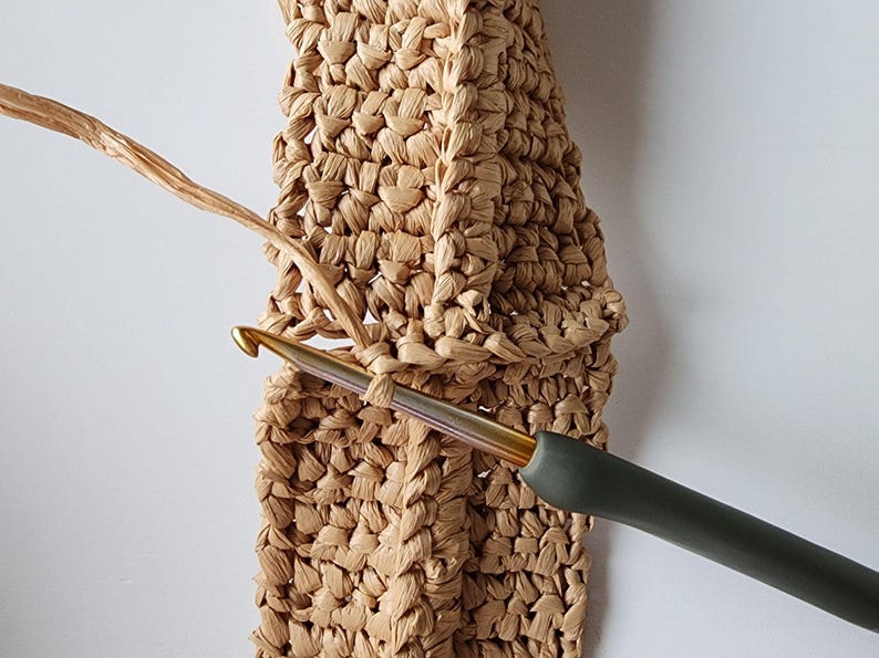 May include: Close-up of a crochet project in progress. A beige woven material is being worked with a gold-colored crochet hook with a dark green handle. The hook is inserted into a loop of the woven material.