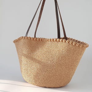 K&ouml;nnte beinhalten: Eine gro&szlig;e, beige geh&auml;kelte Tragetasche mit braunen Lederriemen. Die Tasche hat einen gewellten Rand und ist aus einem gewebten Material gefertigt.