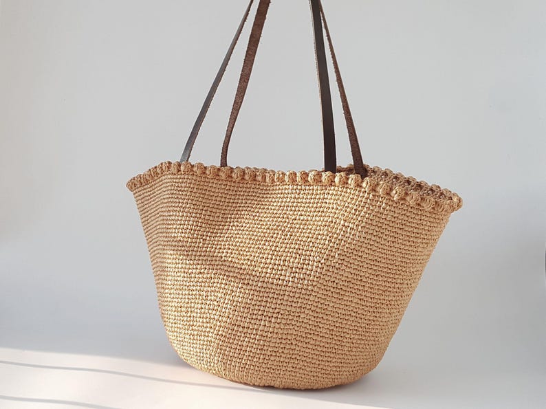 K&ouml;nnte beinhalten: Eine gro&szlig;e, beige geh&auml;kelte Tragetasche mit braunen Lederriemen. Die Tasche hat einen gewellten Rand und ist aus einem gewebten Material gefertigt.