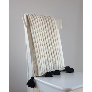 Puede incluir: Manta de punto blanco con borlas negras drapeada sobre una silla blanca.
