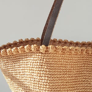 K&ouml;nnte beinhalten: Eine beige geh&auml;kelte Tragetasche mit einem braunen Ledergriff. Die Tasche hat einen gewellten Rand und eine gewebte Textur.