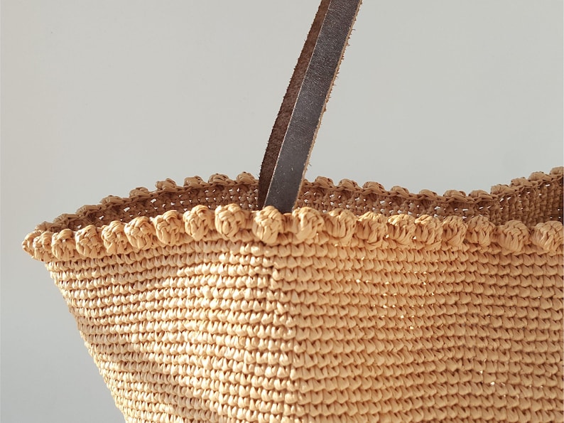 K&ouml;nnte beinhalten: Eine beige geh&auml;kelte Tragetasche mit einem braunen Ledergriff. Die Tasche hat einen gewellten Rand und eine gewebte Textur.