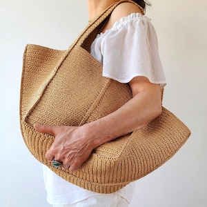 May include: A large, open-top tote bag in a light brown woven material. The bag has a rectangular shape with a wide, flat base and a textured surface. The bag is held by a person wearing a white top.