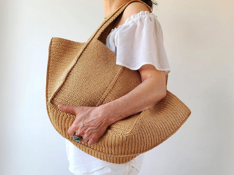 May include: A large, open-top tote bag in a light brown woven material. The bag has a rectangular shape with a wide, flat base and a textured surface. The bag is held by a person wearing a white top.