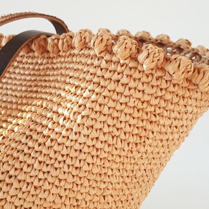 K&ouml;nnte beinhalten: Nahaufnahme einer beige geflochtenen Strohtasche mit einem braunen Lederriemen. Die Tasche hat einen dekorativen Besatz am oberen Rand.
