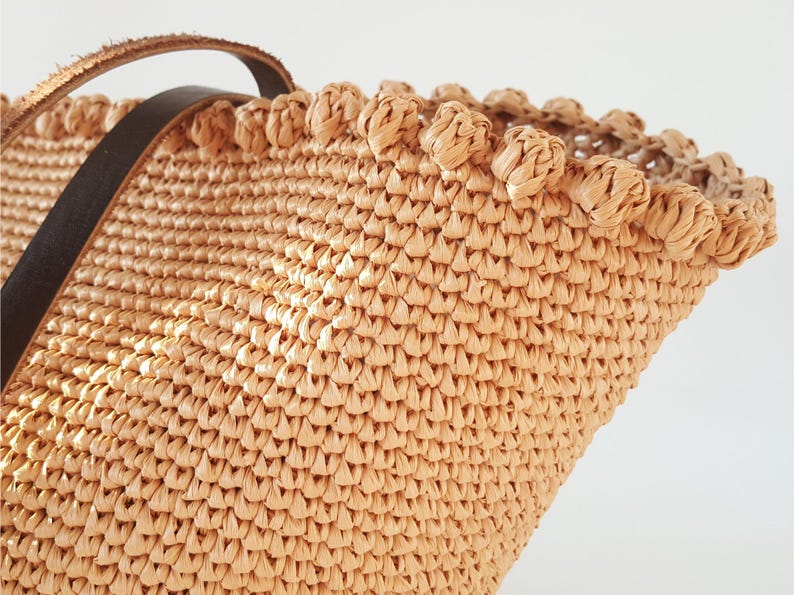 K&ouml;nnte beinhalten: Nahaufnahme einer beige geflochtenen Strohtasche mit einem braunen Lederriemen. Die Tasche hat einen dekorativen Besatz am oberen Rand.