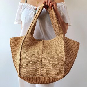May include: A large, beige crochet tote bag with sturdy handles. The bag features a textured stitch pattern and a rectangular base. The bag is held by a person wearing a white top.
