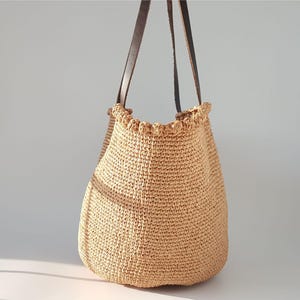 K&ouml;nnte beinhalten: Eine beige geh&auml;kelte Tragetasche mit braunen Lederriemen. Die Tasche hat einen gekr&auml;uselten oberen Rand.