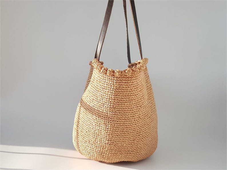 K&ouml;nnte beinhalten: Eine beige geh&auml;kelte Tragetasche mit braunen Lederriemen. Die Tasche hat einen gekr&auml;uselten oberen Rand.