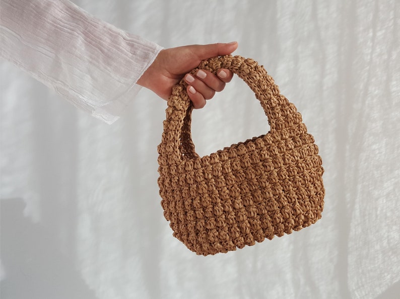 K&ouml;nnte beinhalten: Eine kleine, braune gewebte Handtasche mit einem einzigen Henkel. Die Tasche ist aus einem dicht gewebten stroh&auml;hnlichen Material gefertigt.