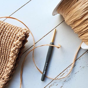 K&ouml;nnte beinhalten: Nahaufnahme einer geh&auml;kelten Tasche aus Naturfasern, einer Spule mit beigefarbenem Raffia-Garn und einer grauen H&auml;kelnadel mit goldener Spitze. Die Nadel ist mit "5,0 mm Clover Amour" beschriftet.