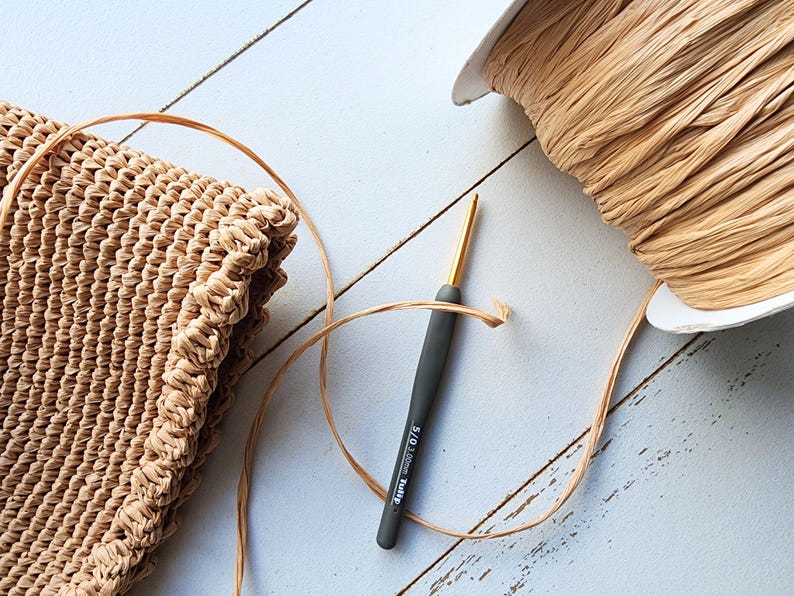 K&ouml;nnte beinhalten: Nahaufnahme einer geh&auml;kelten Tasche aus Naturfasern, einer Spule mit beigefarbenem Raffia-Garn und einer grauen H&auml;kelnadel mit goldener Spitze. Die Nadel ist mit "5,0 mm Clover Amour" beschriftet.