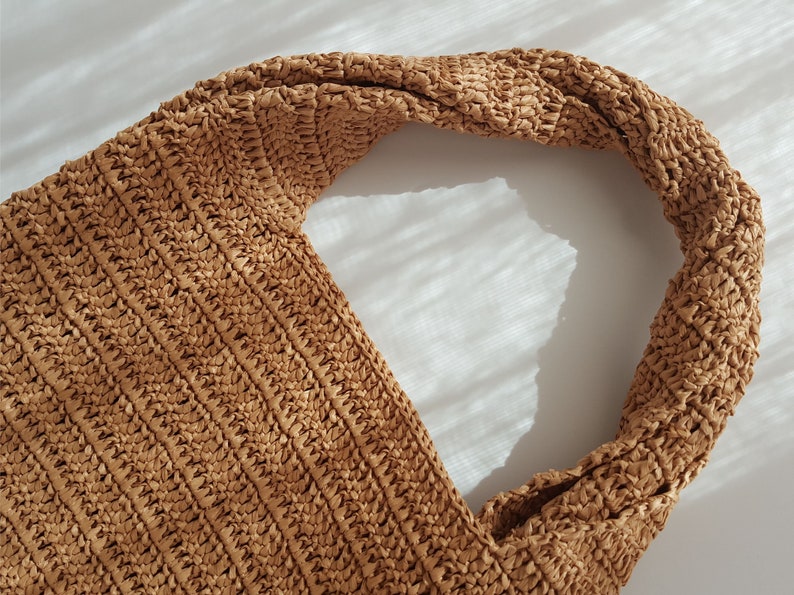 K&ouml;nnte beinhalten: Eine braune gewebte Tragetasche mit einem geknoteten Henkel. Die Tasche ist aus einem strukturierten Material gefertigt und hat einen l&auml;ssigen, entspannten Stil.