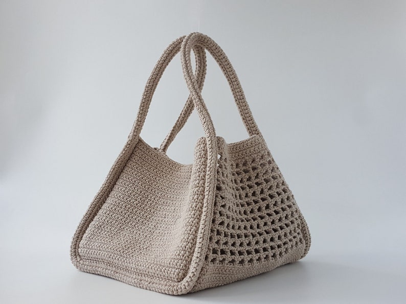 Op de afbeelding: Een beige gehaakte tote bag met een gaasontwerp aan de ene kant en een effen ontwerp aan de andere kant. De tas heeft twee handvatten.