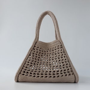 Op de afbeelding: Een beige gehaakte tote bag met een brede, open bovenkant en twee handvatten. De tas is gemaakt van dik, getextureerd garen en heeft een geometrisch patroon.