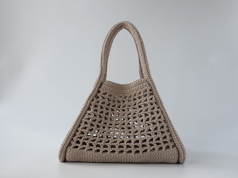 Op de afbeelding: Een beige gehaakte tote bag met een brede, open bovenkant en twee handvatten. De tas is gemaakt van dik, getextureerd garen en heeft een geometrisch patroon.