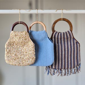 May include: Three handmade crochet handbags hanging from a white rod. The first is a beige bag with gold sequins and a dark wood handle. The second is a solid light blue bag with a light wood handle. The third is a striped bag with fringe.