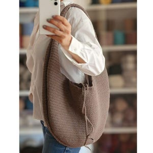 May include: A large, taupe-coloured crochet shoulder bag with a rounded shape. The bag has a thick strap and a drawstring closure. The texture of the crochet is visible, and the bag appears sturdy. The bag is being held.