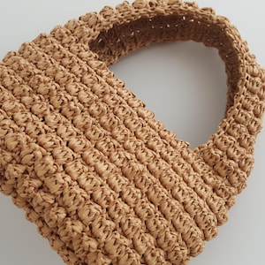 K&ouml;nnte beinhalten: Eine geflochtene Strohtasche in Tan mit abgerundeter Oberseite und einem einzigen Henkel.