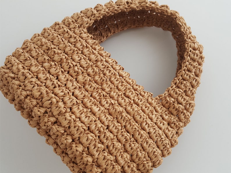 K&ouml;nnte beinhalten: Eine geflochtene Strohtasche in Tan mit abgerundeter Oberseite und einem einzigen Henkel.