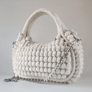 Op de afbeelding: Een witte gehaakte handtas met een zilveren kettingband. De tas heeft een afgeronde vorm en een textuur.