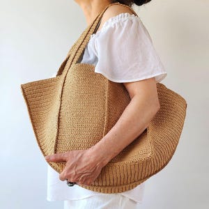 May include: A large, beige crocheted tote bag with sturdy handles. The bag has a rectangular shape with a wide opening and a textured, ribbed pattern. The bag is held by a person wearing a white top.