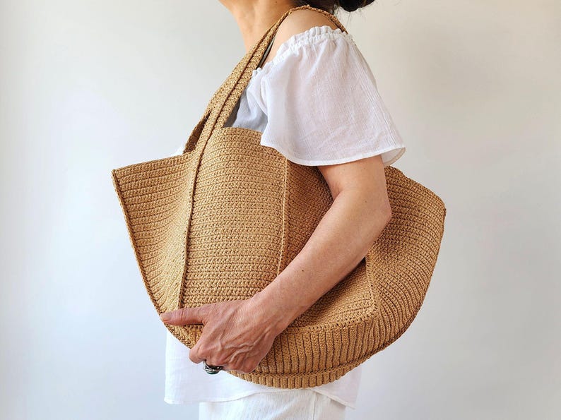 May include: A large, beige crocheted tote bag with sturdy handles. The bag has a rectangular shape with a wide opening and a textured, ribbed pattern. The bag is held by a person wearing a white top.
