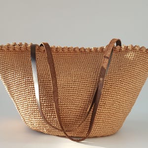 K&ouml;nnte beinhalten: Eine gro&szlig;e, beige geh&auml;kelte Tragetasche mit zwei braunen Lederriemen. Die Tasche hat einen gewellten Rand und eine gewebte Textur.