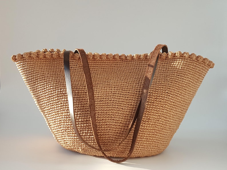 K&ouml;nnte beinhalten: Eine gro&szlig;e, beige geh&auml;kelte Tragetasche mit zwei braunen Lederriemen. Die Tasche hat einen gewellten Rand und eine gewebte Textur.