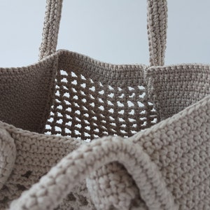 Op de afbeelding: Een beige gehaakte tote bag met een gaasontwerp. De tas heeft twee handvatten en een brede opening.