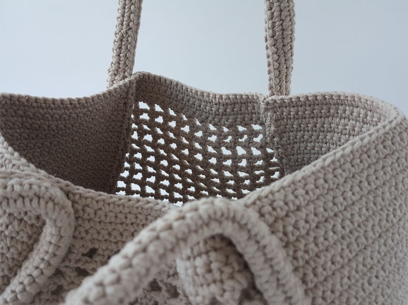 Op de afbeelding: Een beige gehaakte tote bag met een gaasontwerp. De tas heeft twee handvatten en een brede opening.