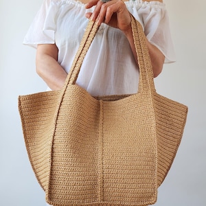 May include: A large, beige crochet tote bag with sturdy handles. The bag features a rectangular base and a wide, open top. The crochet pattern is visible, creating a textured surface. The bag is held by a person wearing a white off-the-shoulder top.