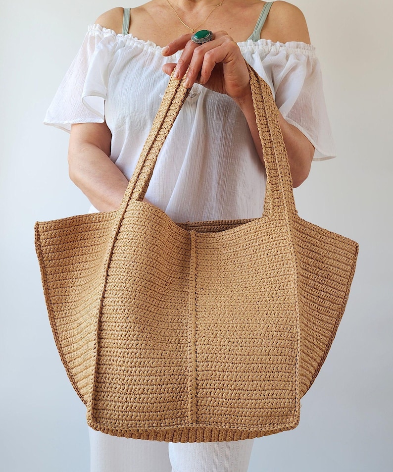 May include: A large, beige crochet tote bag with sturdy handles. The bag features a rectangular base and a wide, open top. The crochet pattern is visible, creating a textured surface. The bag is held by a person wearing a white off-the-shoulder top.