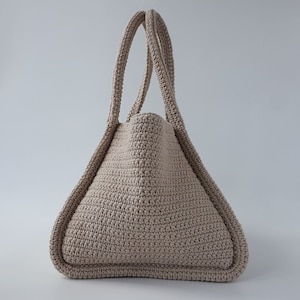 Op de afbeelding: Een beige gehaakte tote bag met twee handvatten. De tas is gemaakt met een textuur-steek en heeft een driehoekige vorm.