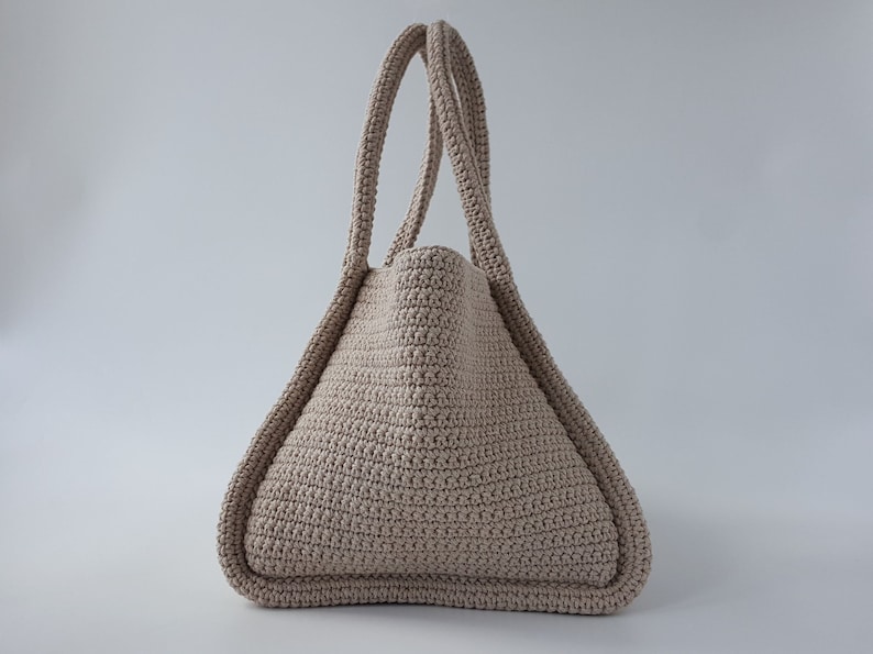 Op de afbeelding: Een beige gehaakte tote bag met twee handvatten. De tas is gemaakt met een textuur-steek en heeft een driehoekige vorm.