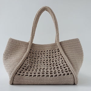 Op de afbeelding: Een beige gehaakte tote bag met een groot open geweven patroon. De tas heeft twee dikke hengsels.