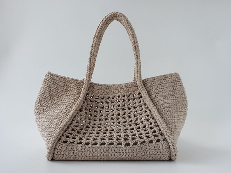 Op de afbeelding: Een beige gehaakte tote bag met een groot open geweven patroon. De tas heeft twee dikke hengsels.